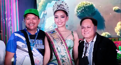 Three individuals at an event, with a woman in the center wearing a crown and sash, flanked by two smiling men, one on each side. The background suggests a celebratory atmosphere, possibly